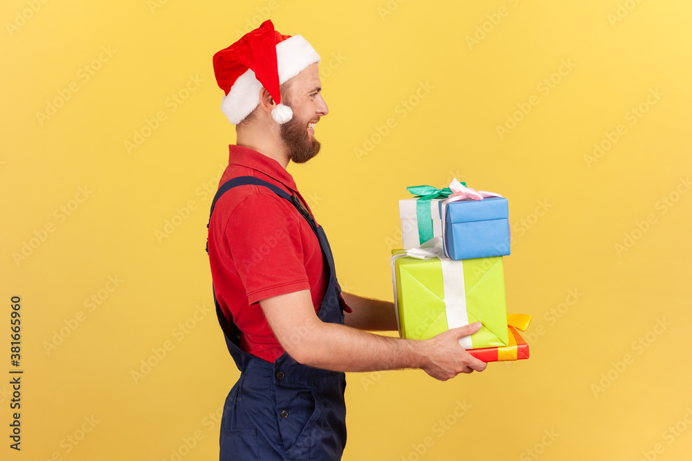 Profile portrait male courier in professional uniform and red santa claus hat holding and giving presents, delivery in holidays. Indoor studio shot isolated on yellow background Profile portrait male courier in professional uniform and red santa claus hat holding and giving presents, delivery in holidays. Indoor studio shot isolated on yellow background