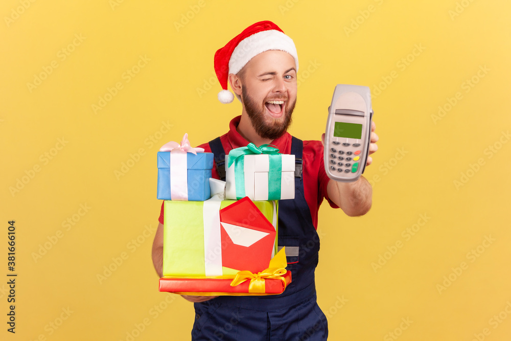 Happy positive courier in santa claus hat holding many gift boxes and showing payment terminal winking at camera, asking to pay contactless. Indoor studio shot isolated on yellow background Happy positive courier in santa claus hat holding many gift boxes and showing payment terminal winking at camera, asking to pay contactless. Indoor studio shot isolated on yellow background