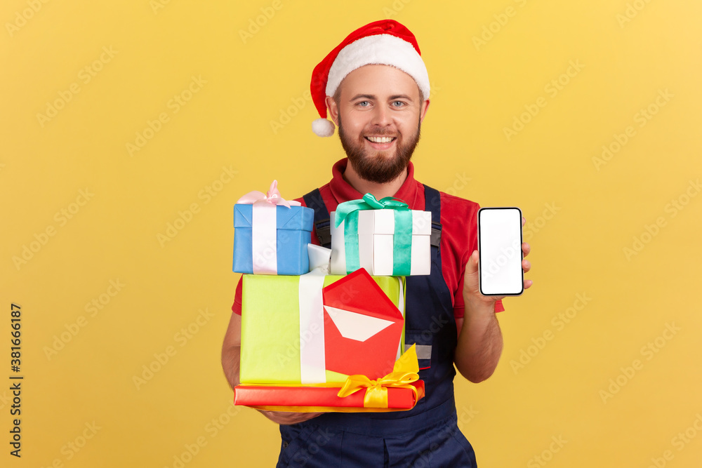 Optimistic deliveryman in uniform and red santa claus hat holding holidays presents and showing smartphone with blank display, order and delivery app. Indoor studio shot isolated on yellow background Optimistic deliveryman in uniform and red santa claus hat holding holidays presents and showing smartphone with blank display, order and delivery app. Indoor studio shot isolated on yellow background