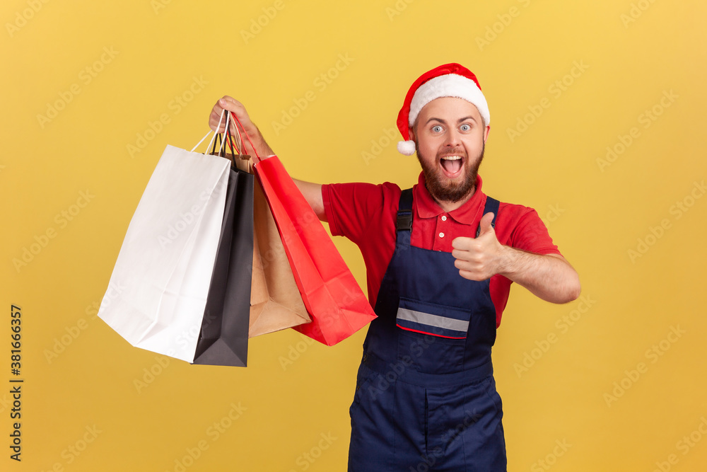Happy excited male courier in uniform and santa claus hat showing thumbs up holding paper shopping bags, likes express delivery. Indoor studio shot isolated on yellow background Happy excited male courier in uniform and santa claus hat showing thumbs up holding paper shopping bags, likes express delivery. Indoor studio shot isolated on yellow background