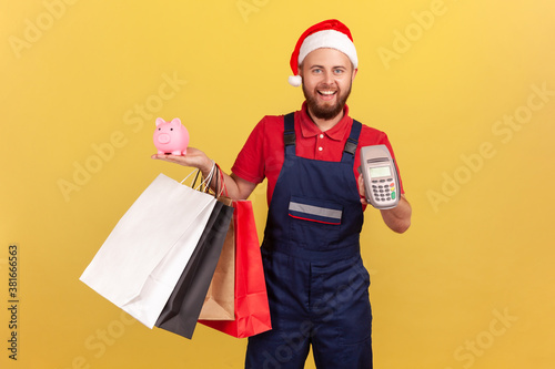 Positive delivery man in uniform and santa claus hat holding paper shopping bags, payment terminal and piggy bank, paying contactless and saving money, cashback. Indoort isolated on yellow background