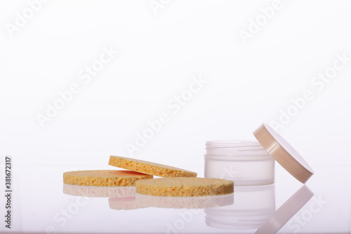 cosmetic cream jar and Three elastic Beige round sponge pads for face make-up cleaning reflected in glass surface isolated on white background. Daily, beauty care cosmetic. Face cream, eye cream
