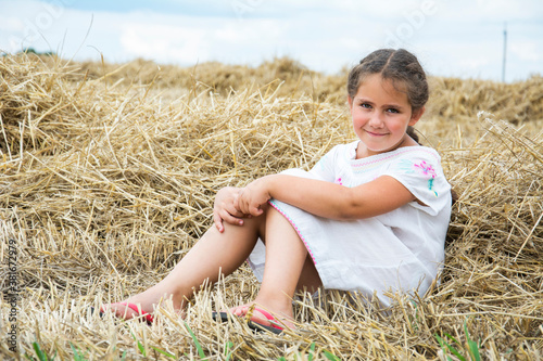 In summer, a little girl is sits on a mown wheat field near the hay.