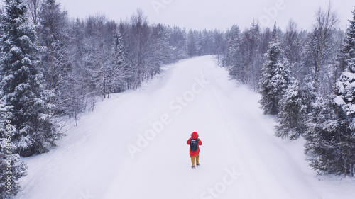 Back view of male tourist in red jacket with rucksack walking on rural path explore wild destination, aerial view of man traveler waiting for car for auto stop during winter, severe weather condition