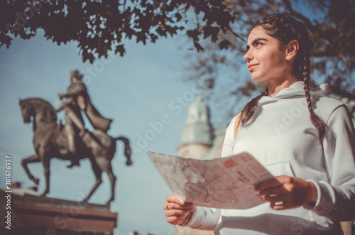 Young cute smiling girl with braids in white hoodie with map in her hands under the tree near the monument on a sightseen tour photo