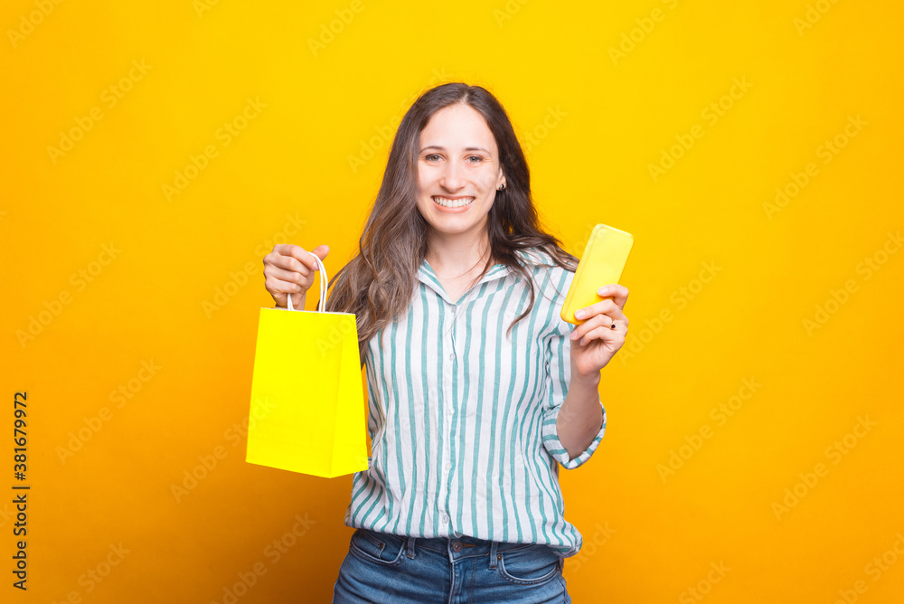 Happy young woman holding a shopping bag and a phone smiling at the camera Happy young woman holding a shopping bag and a phone smiling at the camera