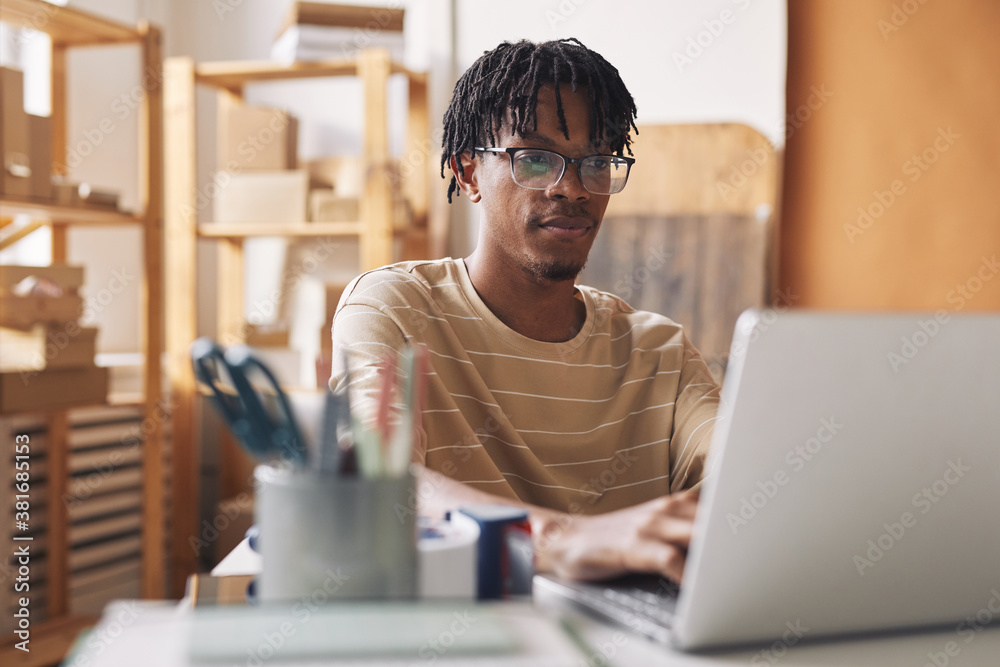 African young man in eyeglasses sitting at the table and working online on laptop at office African young man in eyeglasses sitting at the table and working online on laptop at office