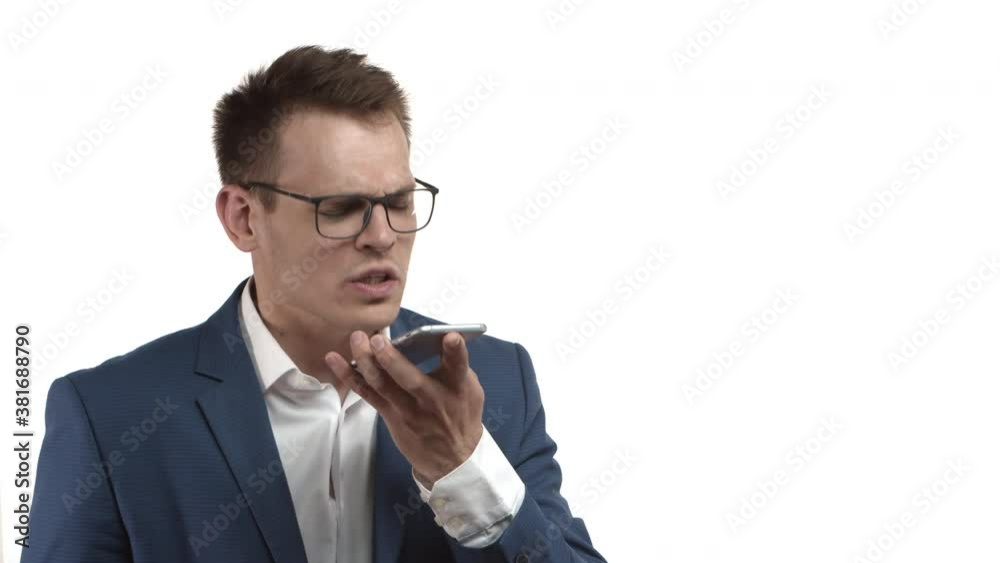 Angry businessman in blue suit and glasses having intense conversation on phone, talking on speakerphone with angry expression, standing over white background Angry businessman in blue suit and glasses having intense conversation on phone, talking on speakerphone with angry expression, standing over white background