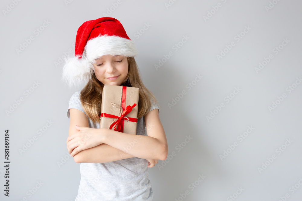 Portrait of a blonde caucasian smiling girl with closed eyes in a Santa hat holding the gift in her hands on a grey background. Christmas and happines concept. Close up, copy space Portrait of a blonde caucasian smiling girl with closed eyes in a Santa hat holding the gift in her hands on a grey background. Christmas and happines concept. Close up, copy space