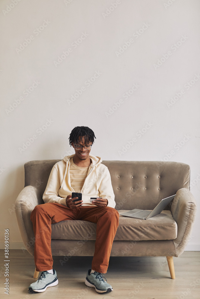African young man sitting on sofa with mobile phone and credit card doing shopping online at home African young man sitting on sofa with mobile phone and credit card doing shopping online at home