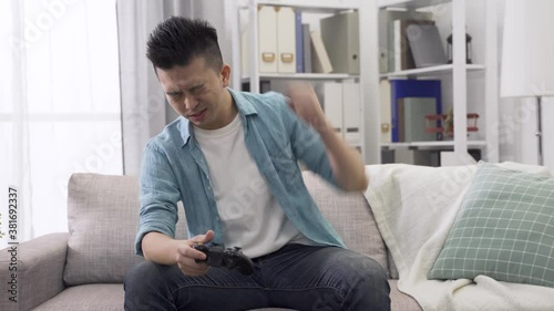 front view asian young guy holding wireless controller auto racing at bright home living room, pounding and patting heavily on lap feeling upset because he lost the game photo