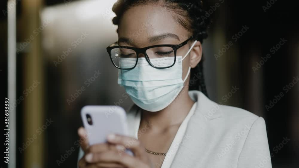 Close-up of young beautiful lady using mobile phone wearing protective mask. Afro-american businesswoman face protection against coronavirus pandemic. Career people. Devices.