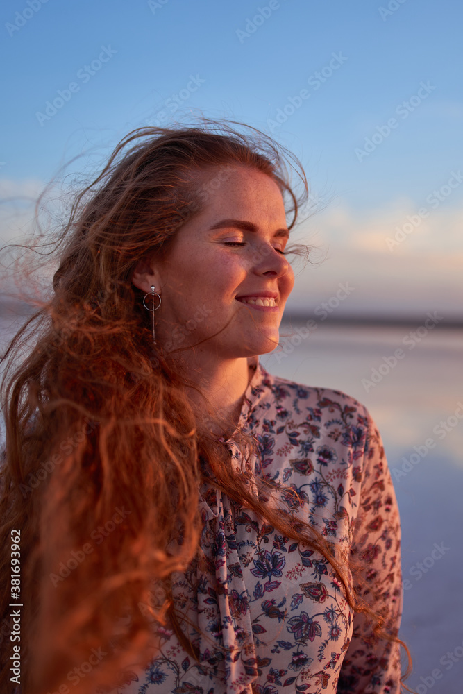Beautiful smiling young woman with flying red hair on the ocean coast at sunset time