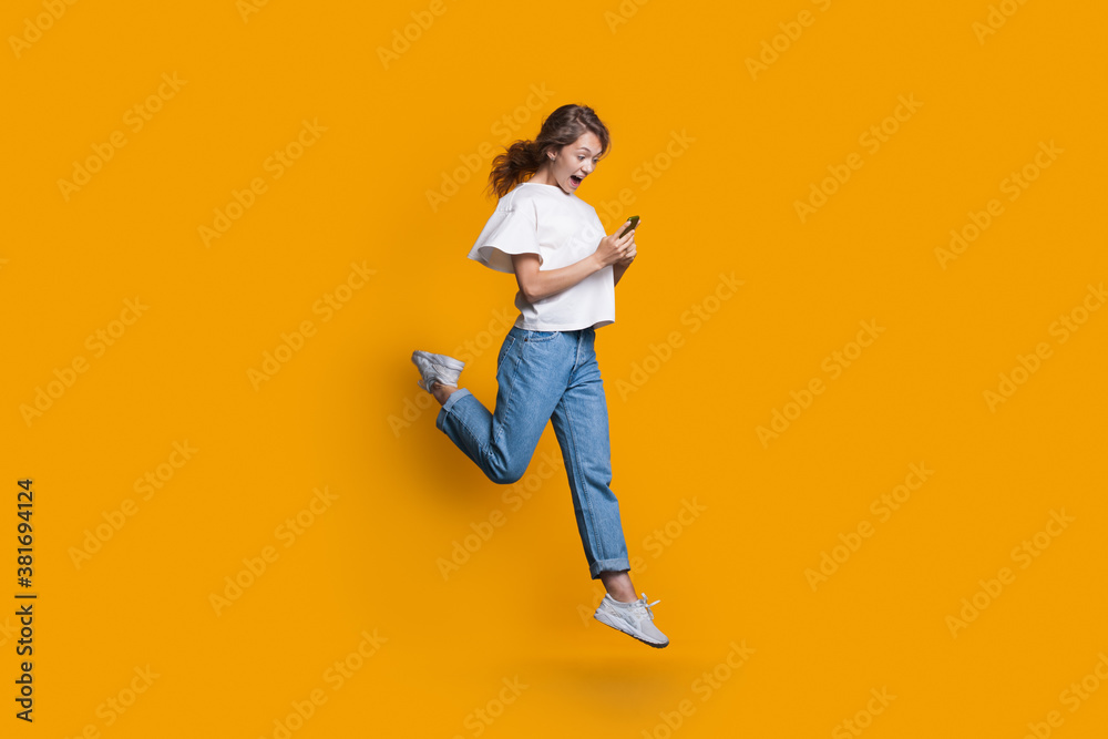 Emotional caucasian blonde woman running and jumping on a yellow studio wall while chatting on mobile and promoting something