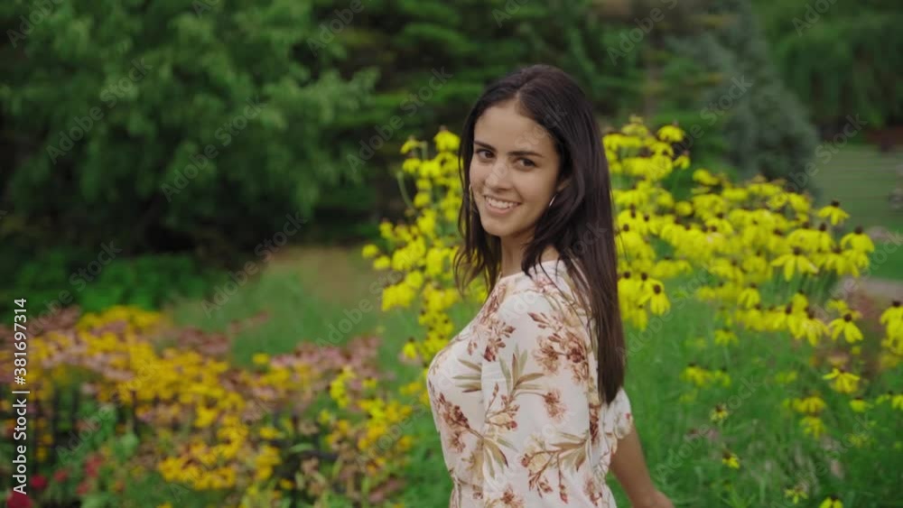 Beautiful brunette woman in a summer dress walking and smiling in a park with flowers on a warm summer day, SLOW MOTION Beautiful brunette woman in a summer dress walking and smiling in a park with flowers on a warm summer day, SLOW MOTION