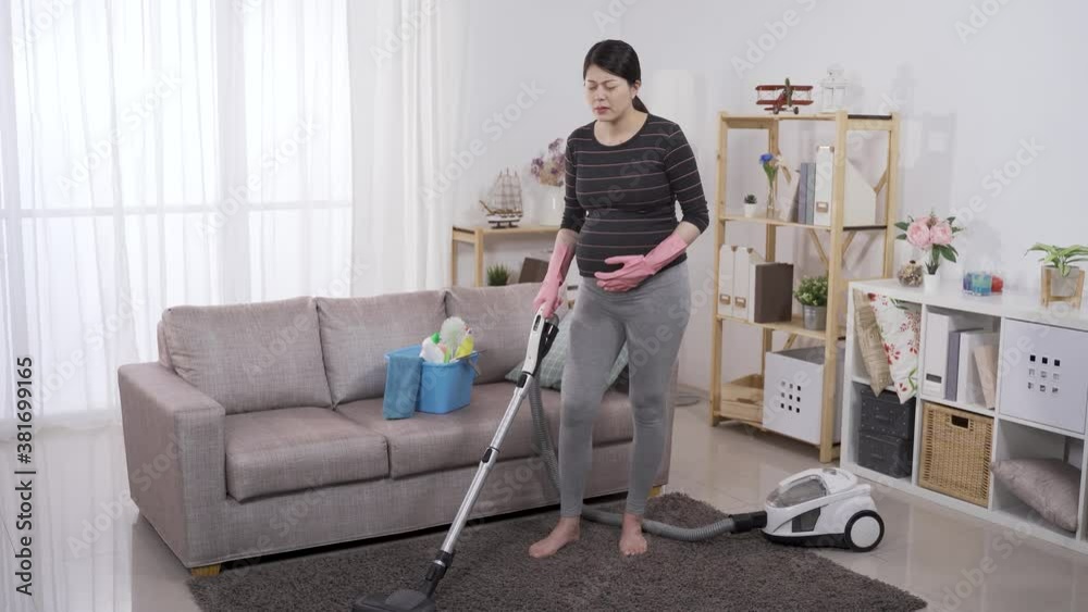 full length asian female in her third trimester feeling sudden painful contraction while cleaning floor with a hoover at home. full length asian female in her third trimester feeling sudden painful contraction while cleaning floor with a hoover at home.