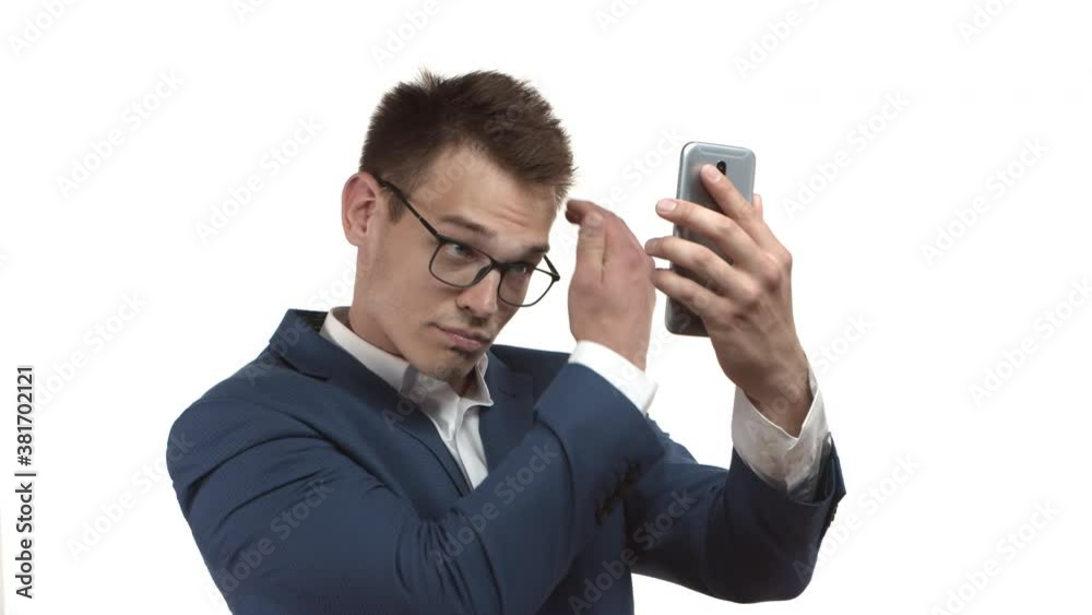 Handsome blond businessman in blue suit and glasses, looking at mobile phone frontal camera to fix and style hair, combing it with hand, standing over white background Handsome blond businessman in blue suit and glasses, looking at mobile phone frontal camera to fix and style hair, combing it with hand, standing over white background