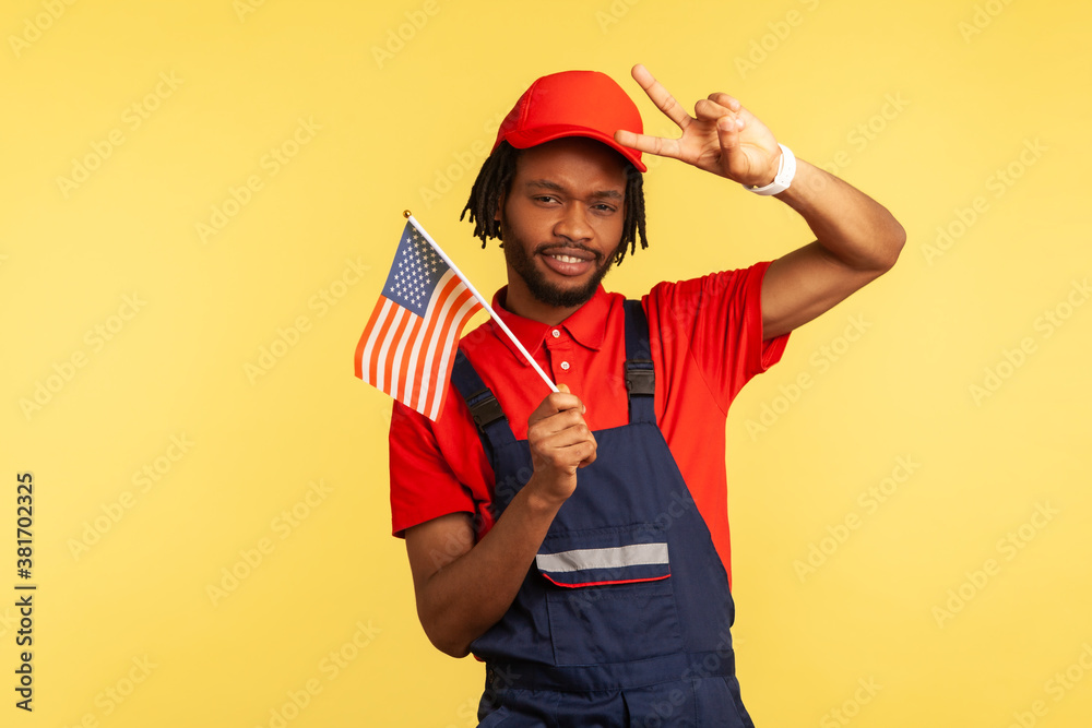 Afro-american male worker in uniform wit beard and dreadlocks holding American flag and showing peace gesture with fingers looking at camera. Indoor studio shot isolated on yellow background Afro-american male worker in uniform wit beard and dreadlocks holding American flag and showing peace gesture with fingers looking at camera. Indoor studio shot isolated on yellow background