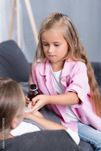 Selective focus of girl giving syrup to sick mother on sofa