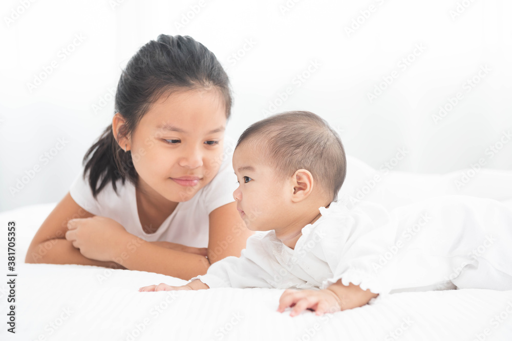 Big asian sister looks with adoration at little brother on white bed. Portrait of a newborn baby with big sister to play at bedroom. Big asian sister looks with adoration at little brother on white bed. Portrait of a newborn baby with big sister to play at bedroom.