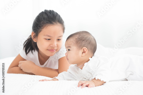 Big asian sister looks with adoration at little brother on white bed. Portrait of a newborn baby with big sister to play at bedroom.