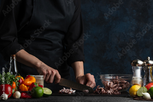 Professional chef cuts octopuses for cooking, on the background of vegetables on a dark blue background. Asian cuisine. Traditional Thai recipe. Seafood concept. Background of preparing dish.