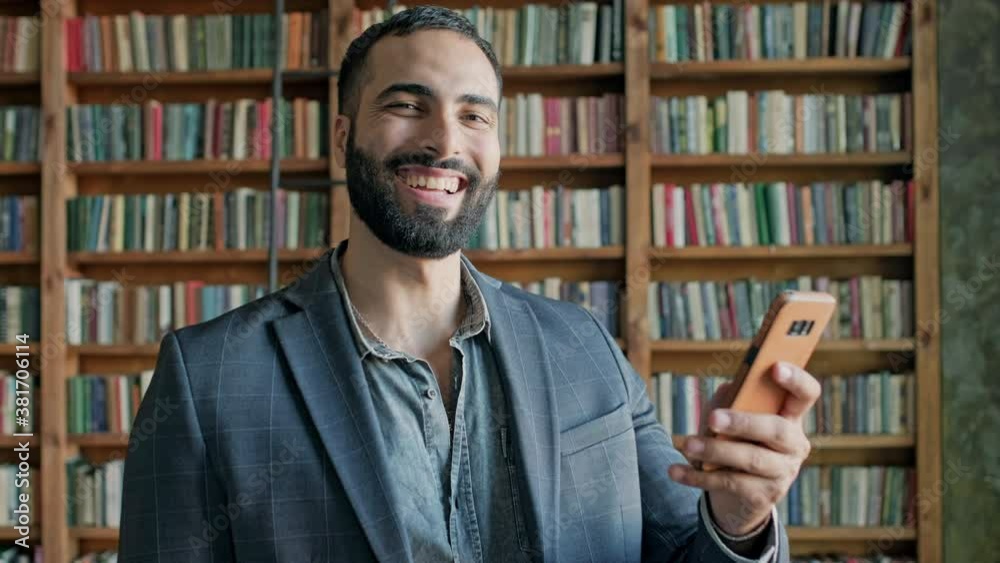 Smiling Young Man In The Library With Mobile Phone In His Hands. Smiling Young Man In The Library With Mobile Phone In His Hands.