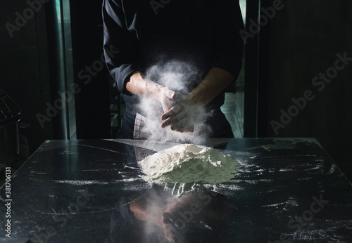 Chef man clap with splash white flour dust man hand on low light dark black background. An experienced chef in a professional kitchen prepares the dough with flour to make bakery.
