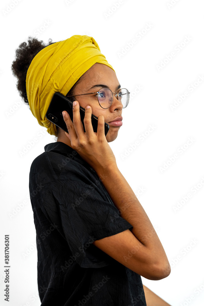 Portrait of young woman wearing glasses and yellow head scarf and speaking on mobile phone over white background