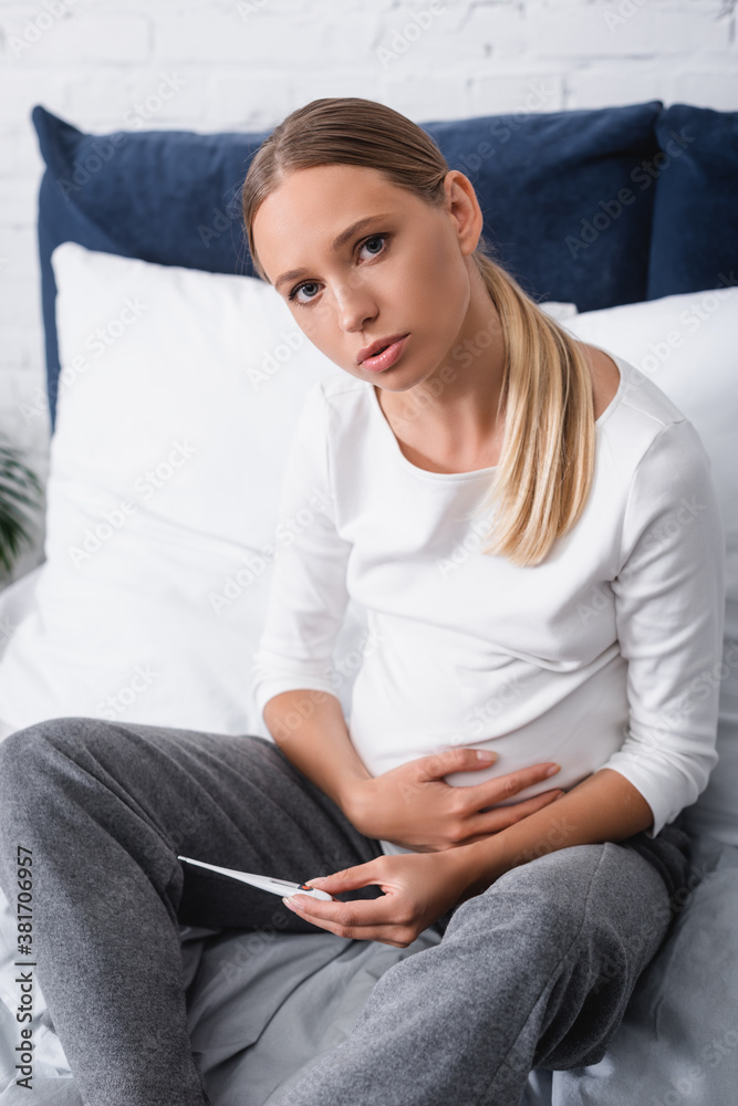 Obraz premium Young pregnant woman looking at camera while holding thermometer in bedroom
