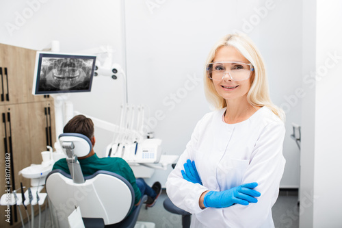 Portrait of a woman dentist smiling, in protective glasses with crossed hands, background dentistry clinic with a patient