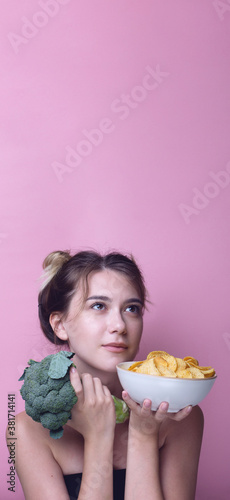 Concept of a choice between a bowl of potato chips and broccoli, portrait of Woman on pink background. junk food , health snack`