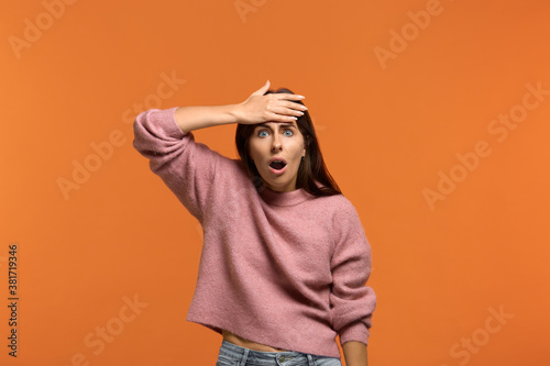 Crap, I forgot to do this. Portrait of worried stunned woman in pink sweater slapping forehead with palm and dropping jaw, recalling important thing and being troubled, on orange background © 5M
