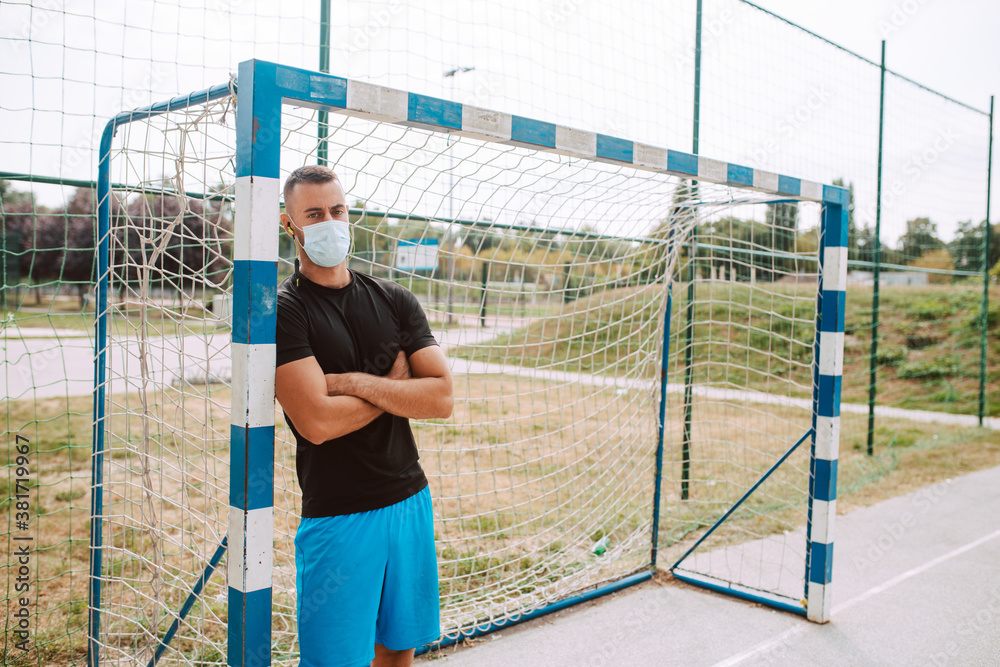 A portrait of a young muscular caucasian male athlete with a face mask standing next to a goal on the football field. COVID - 19 coronavirus protection
