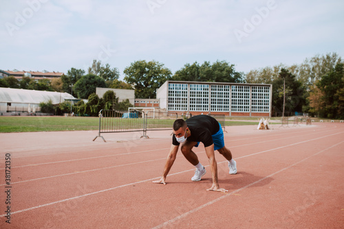 Fit young male caucasian athlete with face mask starting his sprint on an all-weather running track. Muscular athlete outdoors. COVID - 19 coronavirus protection