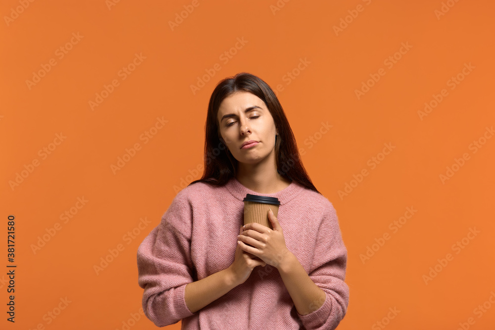 Obraz premium Need to cheer up. Tired sleepy beautiful woman in pink sweater with a tortured look has sad expression, closes eyes, keeps disposable cup of drink containing caffeine. on orange background