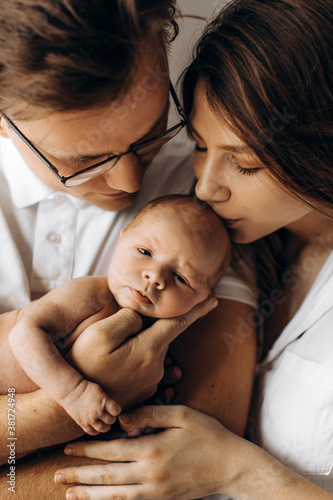 Happy parents with cute newborn baby girl, handsome dad hold little daughter in arms, beautiful wife and loving husband gently kiss their child, young family concept