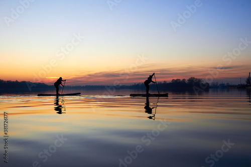 Silhouettes of two women paddle on stand up paddle boarding (SUP) on quiet winter or autumn Danube river at sunset. Colorful sunset over the river with silhouettes of people