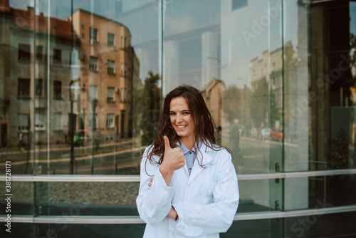 A young female doctor stands in front of the clinic and gives a thumbs up. Doctor portrait during the COVID-19 coronavirus pandemic