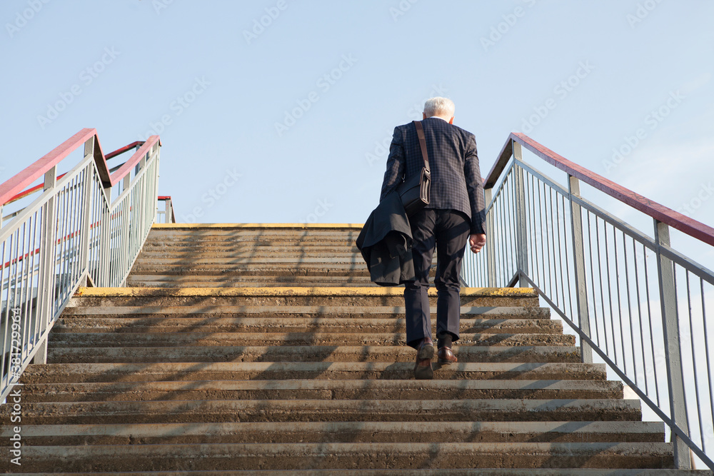 A gray-haired, elderly, well-dressed alone man, wearing a jacket, with a leather bag on his shoulder, climbs up the stairs at the train station. Rear view, no face visible.