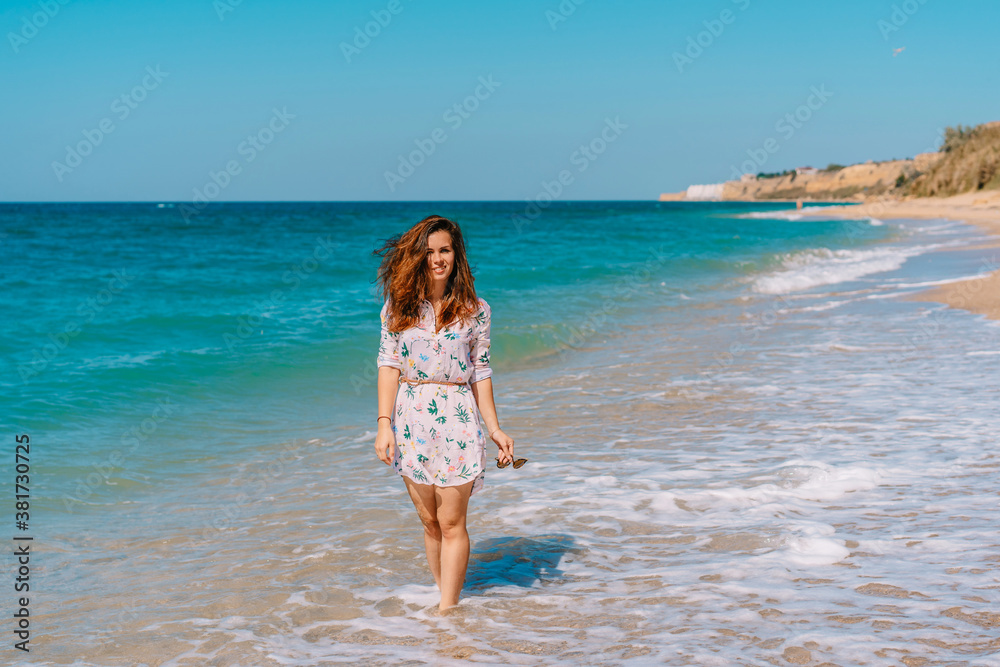 Beautiful smiling woman in a dress walking on the sand along the emerald sea, leisure and tourism concept Beautiful smiling woman in a dress walking on the sand along the emerald sea, leisure and tourism concept