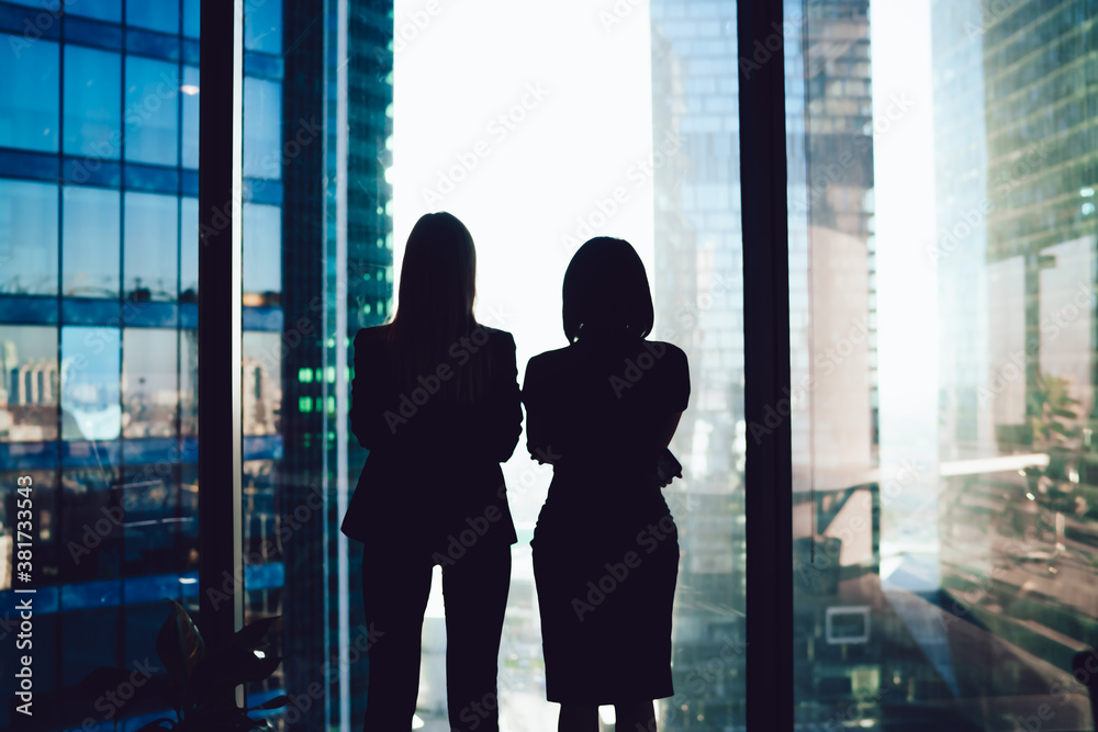 Back view of female colleagues in formal wear standing near window looking at modern exterior of skyscrapers in business center, silhouette of women together planning future success of brainstorming Back view of female colleagues in formal wear standing near window looking at modern exterior of skyscrapers in business center, silhouette of women together planning future success of brainstorming