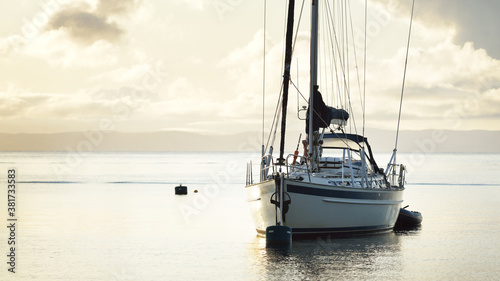 Tableau sur toile A two-masted sailboat (ketch) with a motorboat anchored on mooring near the shore of Craighouse at sunset