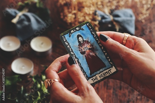 Closeup of wiccan witch holding a small hand painted (made up) Hermit tarot card in her hands. Cozy warm Yule winter solstice (Christmas) background with dried flowers, ivy, holy plants, three candles