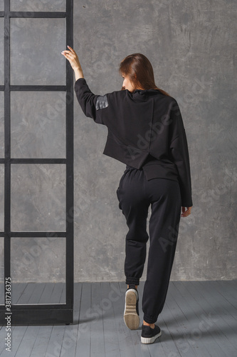 Young woman in black sportswear, pants and sweatshirt. Back view. Concept of fashionable sport outfit, indoors photo.
