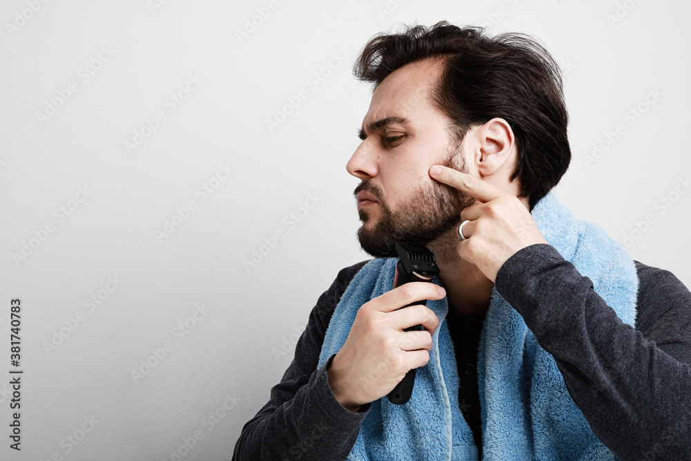 Portrait of handsome young man shaving his beard in bathroom, holding shaver machine. Space for text. Portrait of handsome young man shaving his beard in bathroom, holding shaver machine. Space for text.