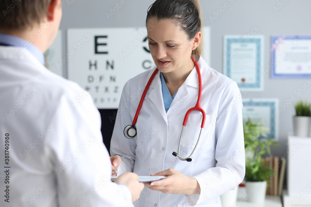 Young woman doctor signs document in hand of another doctor in office in clinic. Collaboration in medicine consultation concept. Young woman doctor signs document in hand of another doctor in office in clinic. Collaboration in medicine consultation concept.