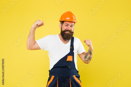 Power building. Strong construction man flex arms yellow background. Bearded man wear hard hat and work uniform. Renovation and repair. Constructing and engineering