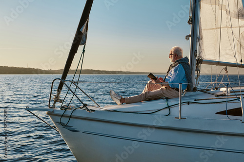 Luxury lifestyle. Side view of a relaxed senior man sitting on the side of sailboat or yacht floating in the calm blue sea at sunset and enjoying amazing view, using digital tablet