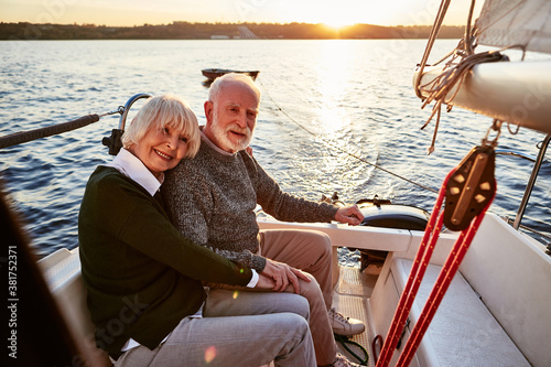 Happy senior couple in love, elderly man and woman holding hands, hugging and spending time together while sitting on the side of yacht deck floating in sea on a sunny day